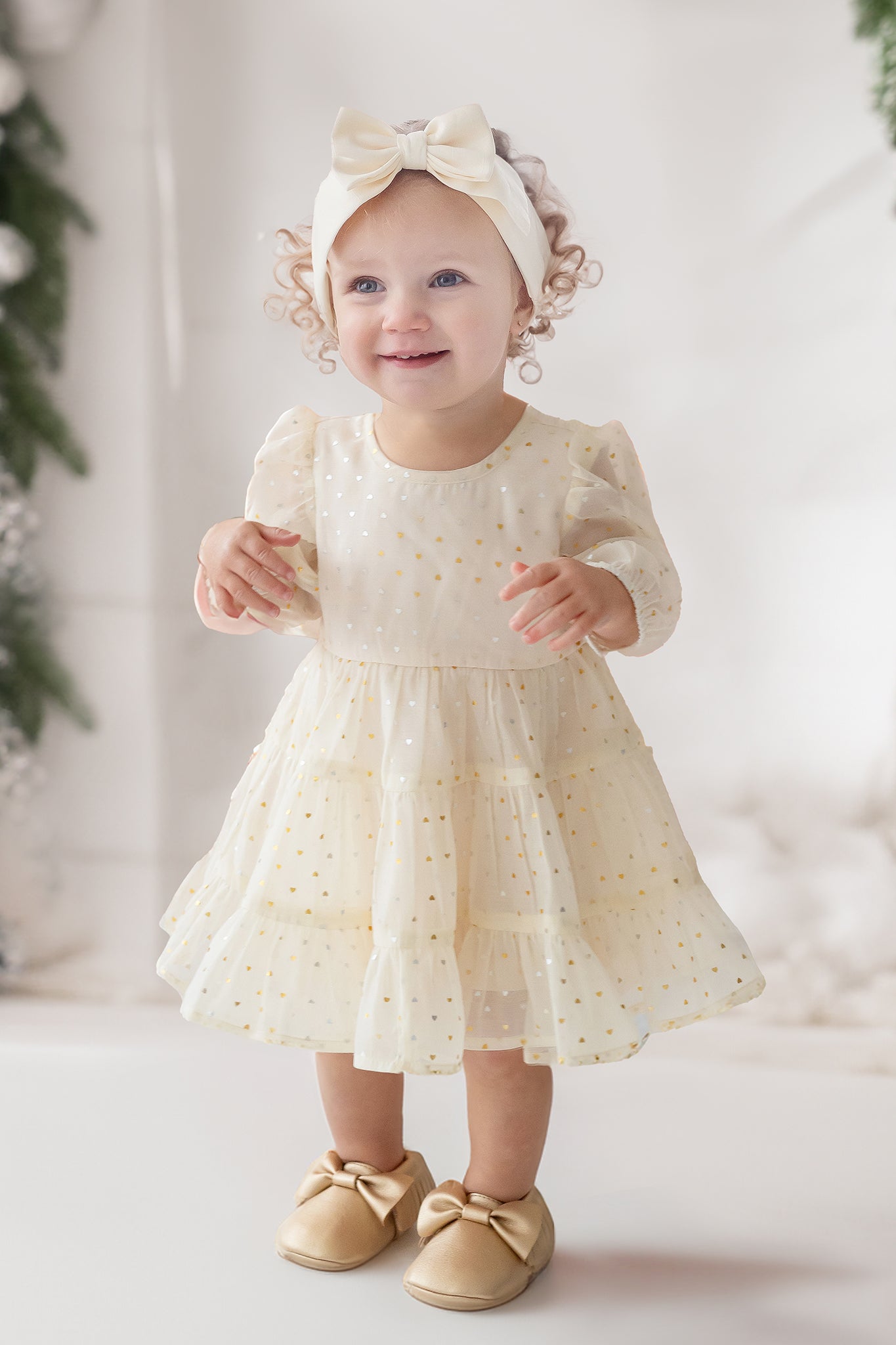 A young toddler smiles indoors wearing the Isobella and Chloe Heart of Gold Little Big Girl Dress—an ivory tulle dress with a layered ruffle skirt—paired with bow shoes and a headband, surrounded by greenery and white decor.