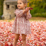 A young girl wears the Mabel and Honey Autumn Floral Dress Long Sleeve as she stands on fallen autumn leaves, smiling and looking to the side while holding red leaves. Blurred trees and a stone structure are in the background.