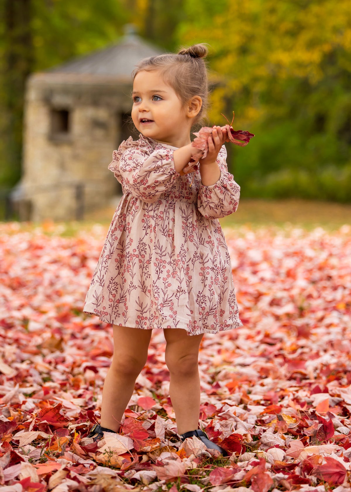 A young girl wears the Mabel and Honey Autumn Floral Dress Long Sleeve as she stands on fallen autumn leaves, smiling and looking to the side while holding red leaves. Blurred trees and a stone structure are in the background.