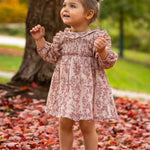 A smiling toddler wears the Mabel and Honey Autumn Floral Dress Long Sleeve, standing on red autumn leaves near a tree, with green grass and blurred trees in the background.