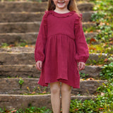 A young girl with long brown hair smiles brightly outdoors, wearing the Mabel and Honey Autumn Dress Red with ruffle details, surrounded by greenery and fallen leaves on stone steps.