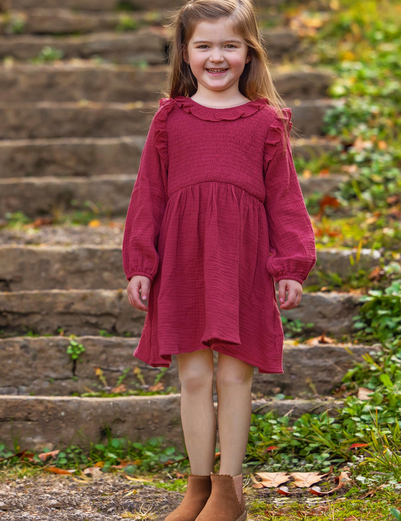 A young girl with long brown hair smiles brightly outdoors, wearing the Mabel and Honey Autumn Dress Red with ruffle details, surrounded by greenery and fallen leaves on stone steps.