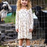 A smiling young girl stands in front of a wire fence with two goats behind her, wearing the Mabel and Honey Drama Llama Dress, paired with white socks and black boots.