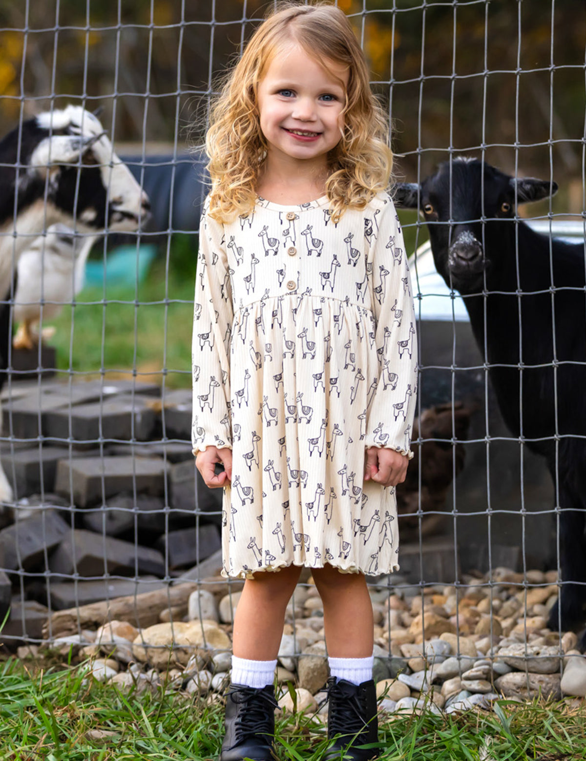 A smiling young girl stands in front of a wire fence with two goats behind her, wearing the Mabel and Honey Drama Llama Dress, paired with white socks and black boots.