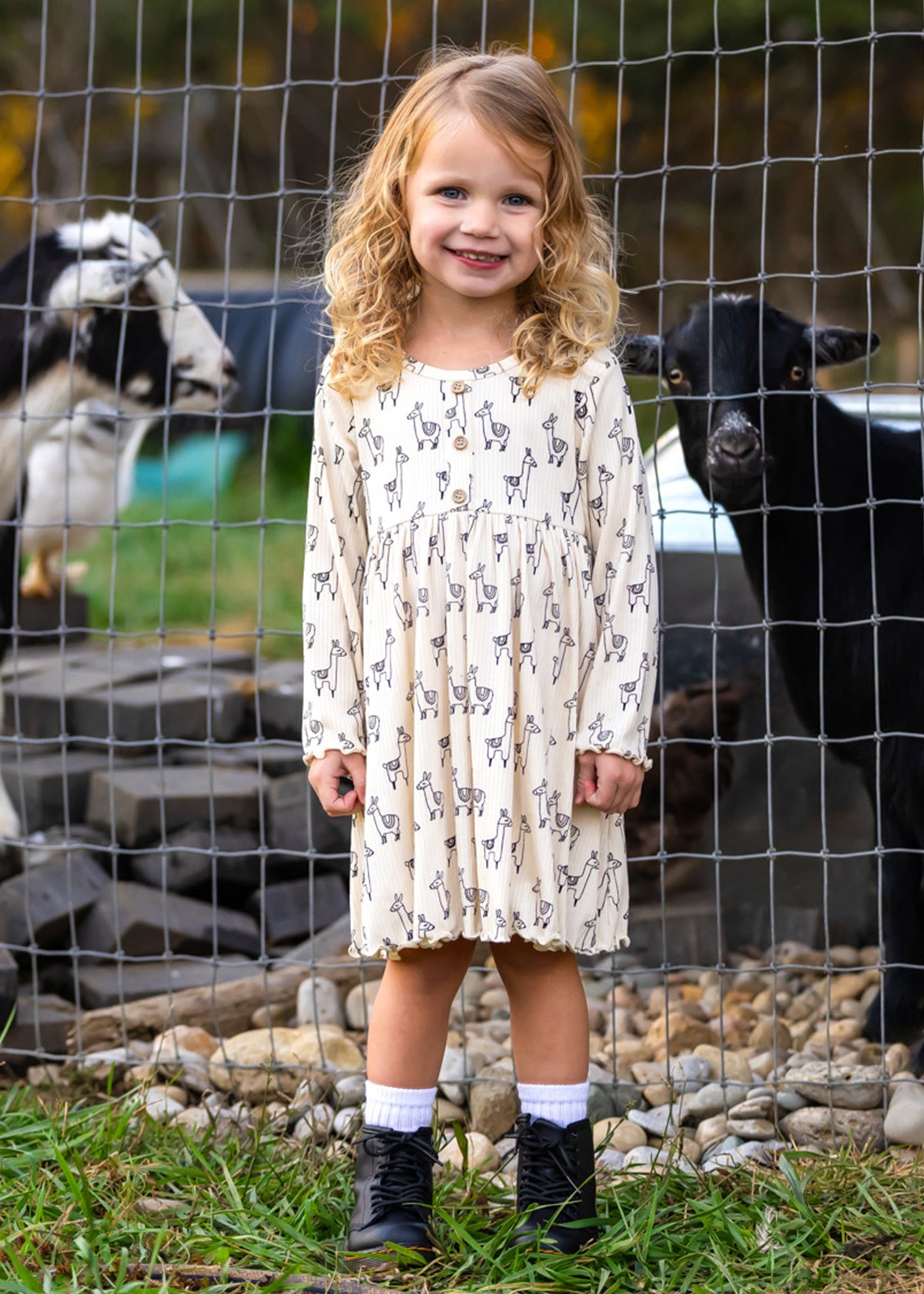 A smiling young girl stands in front of a wire fence with two goats behind her, wearing the Mabel and Honey Drama Llama Dress, paired with white socks and black boots.