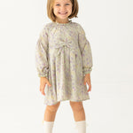 A young girl with light brown hair smiles in the Mabel and Honey Lavender Meadows Floral Bow Dress, featuring a statement bow on the front. She pairs it with white socks and brown shoes, standing against a plain white background.