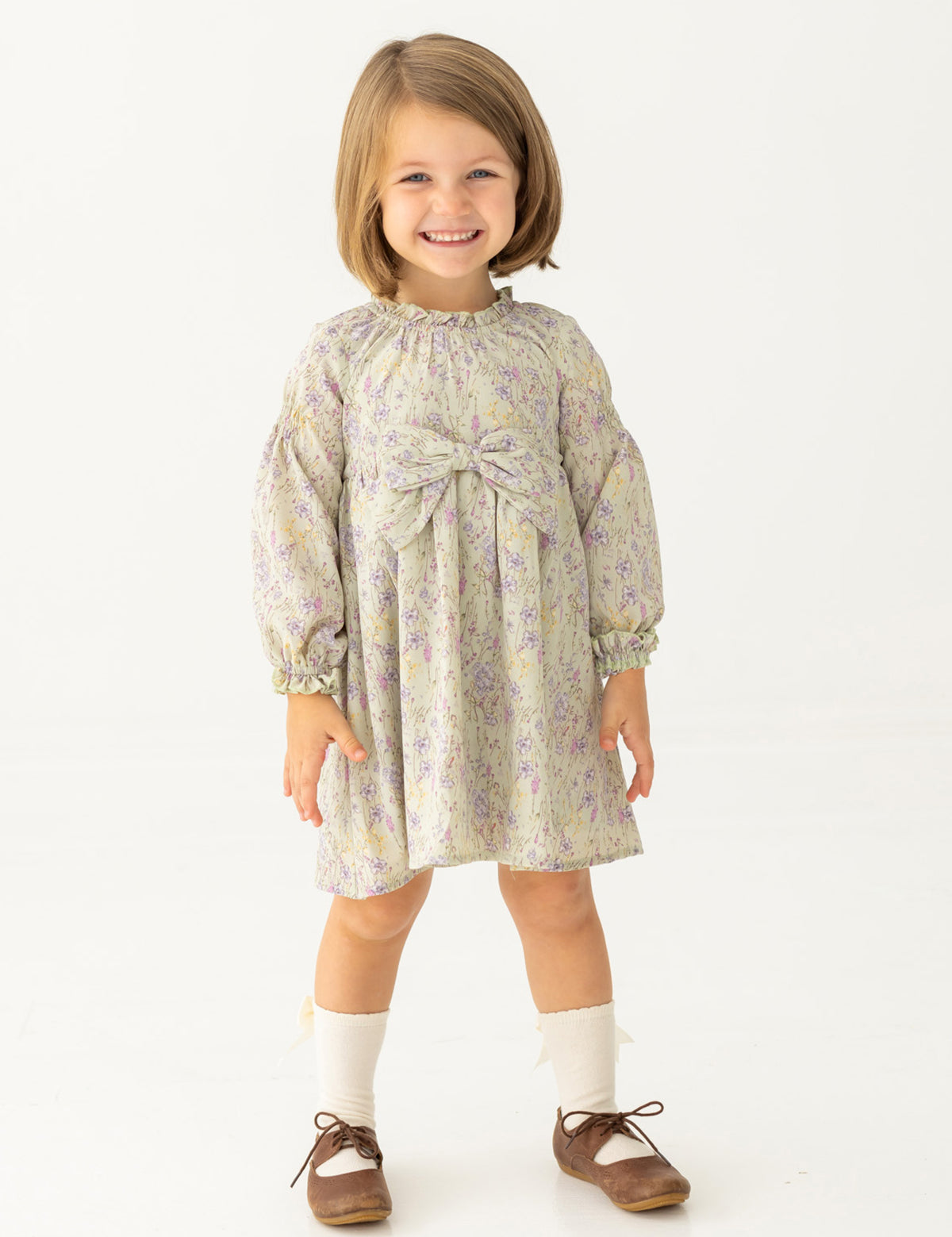 A young girl with light brown hair smiles in the Mabel and Honey Lavender Meadows Floral Bow Dress, featuring a statement bow on the front. She pairs it with white socks and brown shoes, standing against a plain white background.