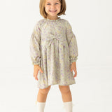 A young girl with light brown hair smiles in the Mabel and Honey Lavender Meadows Floral Bow Dress, featuring a statement bow on the front. She pairs it with white socks and brown shoes, standing against a plain white background.