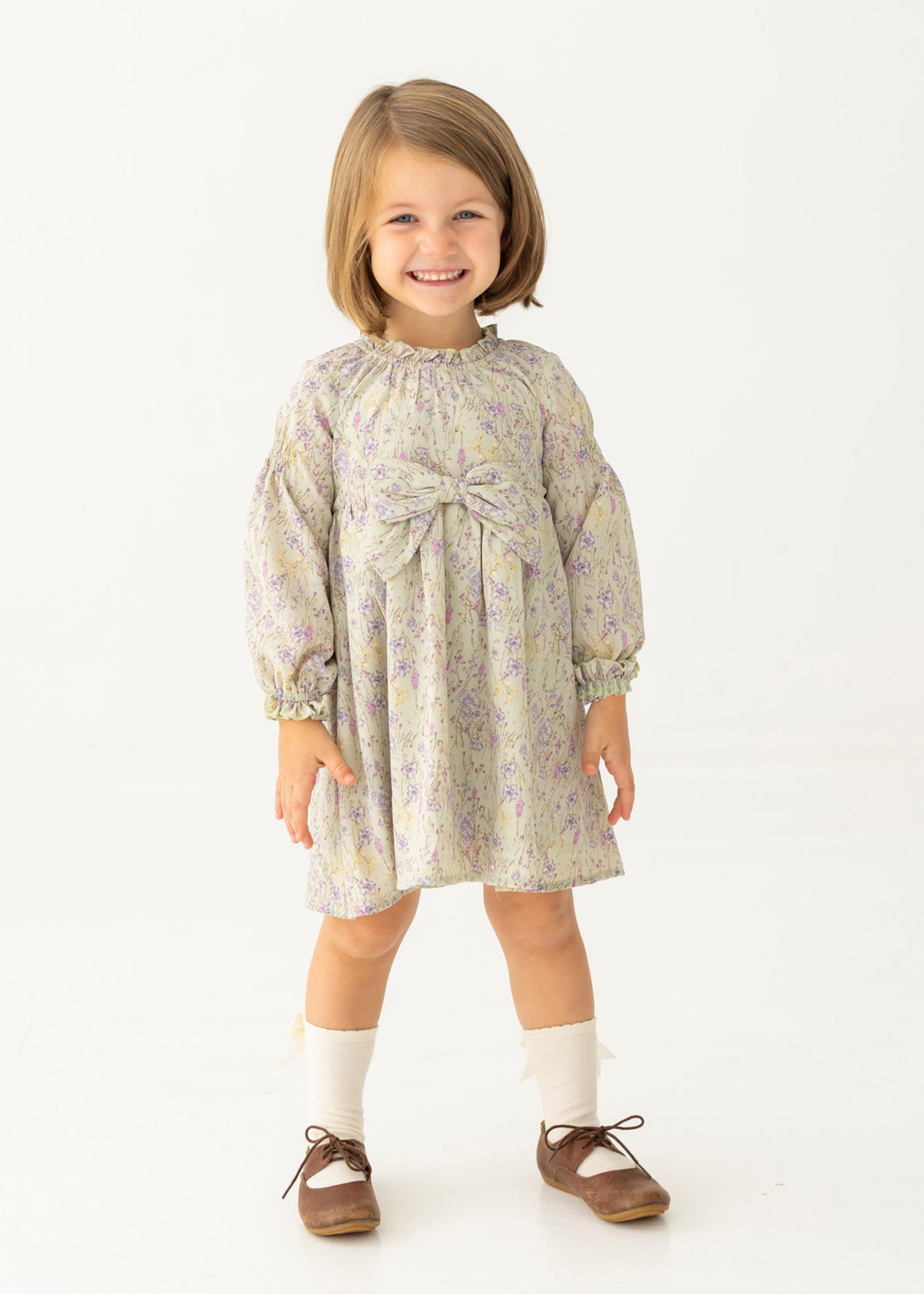 A young girl with light brown hair smiles in the Mabel and Honey Lavender Meadows Floral Bow Dress, featuring a statement bow on the front. She pairs it with white socks and brown shoes, standing against a plain white background.