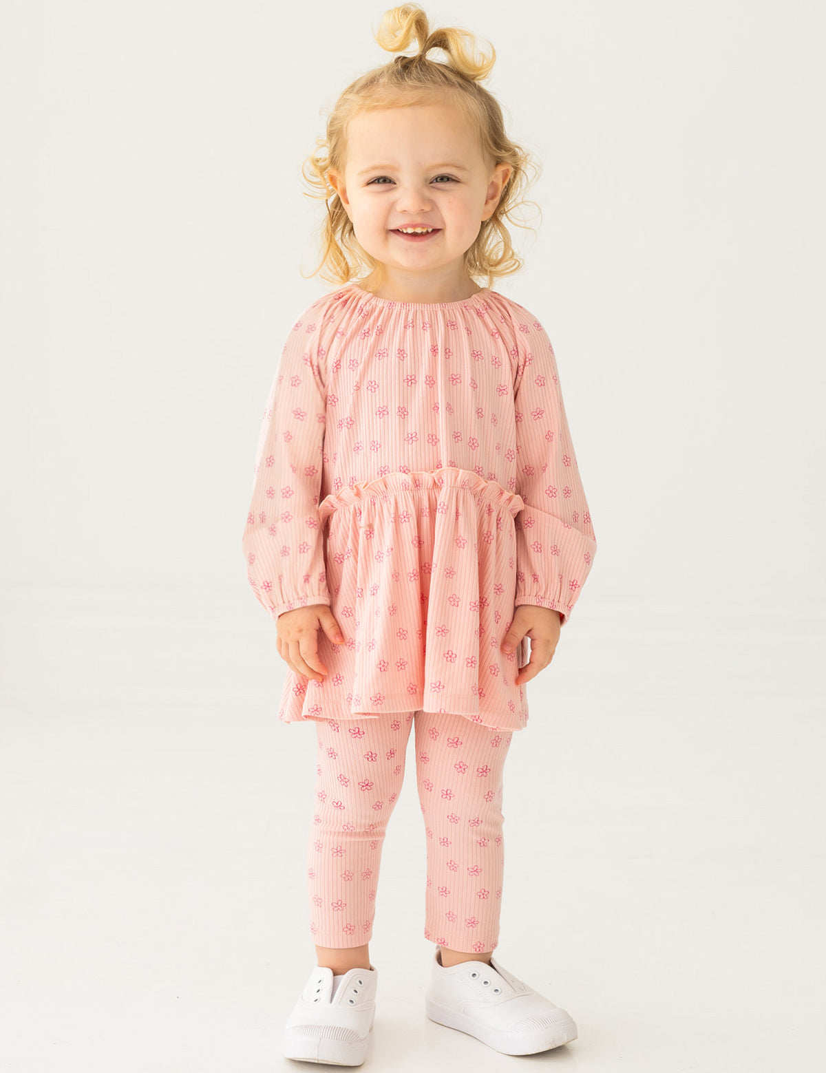 A smiling toddler with blonde hair in a top knot models the Mabel and Honey Dainty Doodle Two Piece Set, featuring a light pink peplum top and matching leggings, paired with white sneakers, against a plain white background.