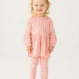A smiling toddler with blonde hair in a top knot models the Mabel and Honey Dainty Doodle Two Piece Set, featuring a light pink peplum top and matching leggings, paired with white sneakers, against a plain white background.