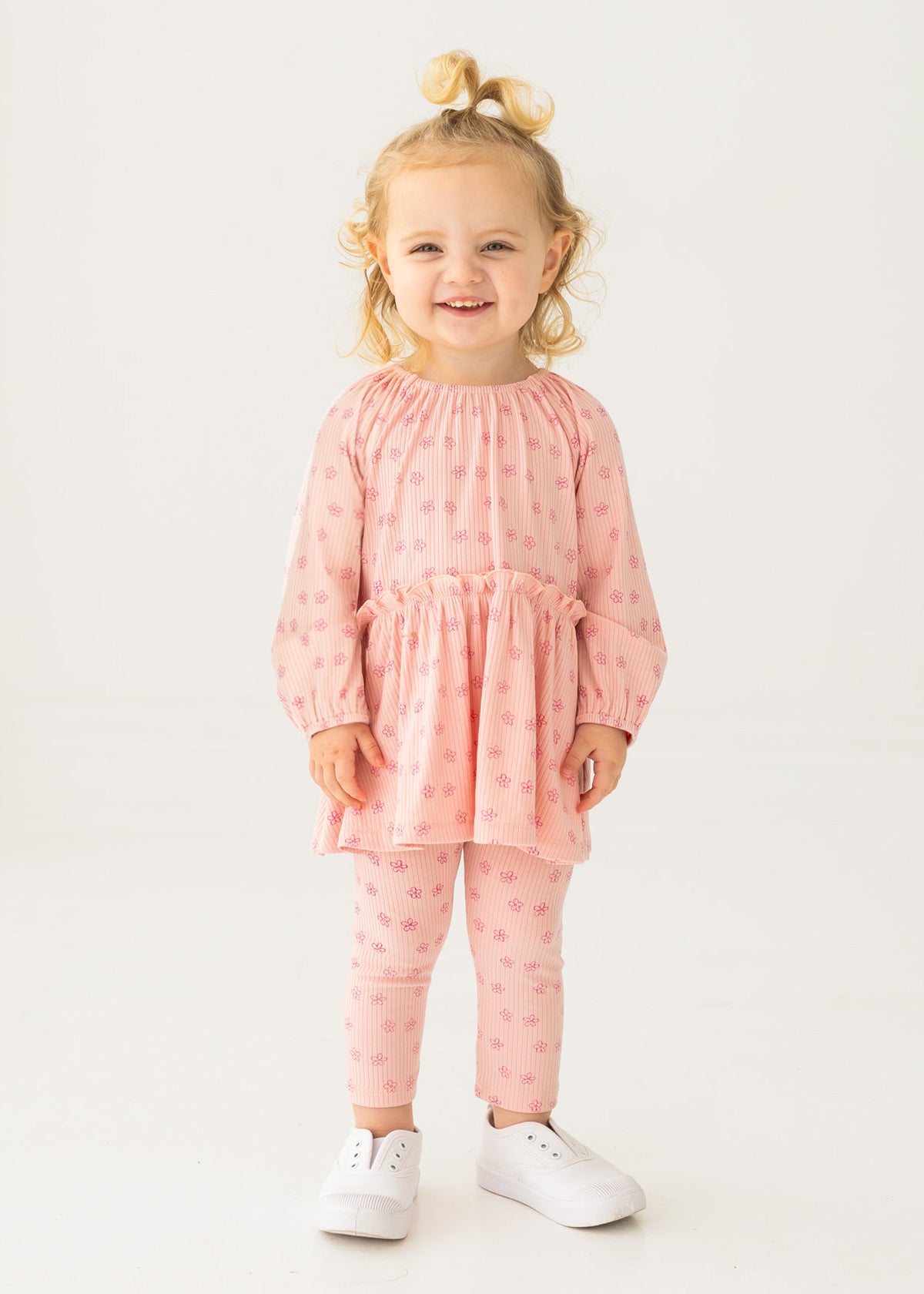 A smiling toddler with blonde hair in a top knot models the Mabel and Honey Dainty Doodle Two Piece Set, featuring a light pink peplum top and matching leggings, paired with white sneakers, against a plain white background.
