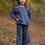 A young girl smiles outdoors on a leaf-strewn path, wearing the Mabel and Honey Robyn Two Piece Set with a ruffled peplum top, wide-leg pants, white shoes, and a big blue hair bow. Fall leaves blur in the background.