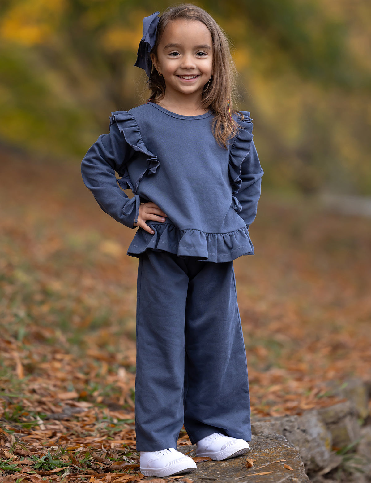 A young girl smiles outdoors on a leaf-strewn path, wearing the Mabel and Honey Robyn Two Piece Set with a ruffled peplum top, wide-leg pants, white shoes, and a big blue hair bow. Fall leaves blur in the background.