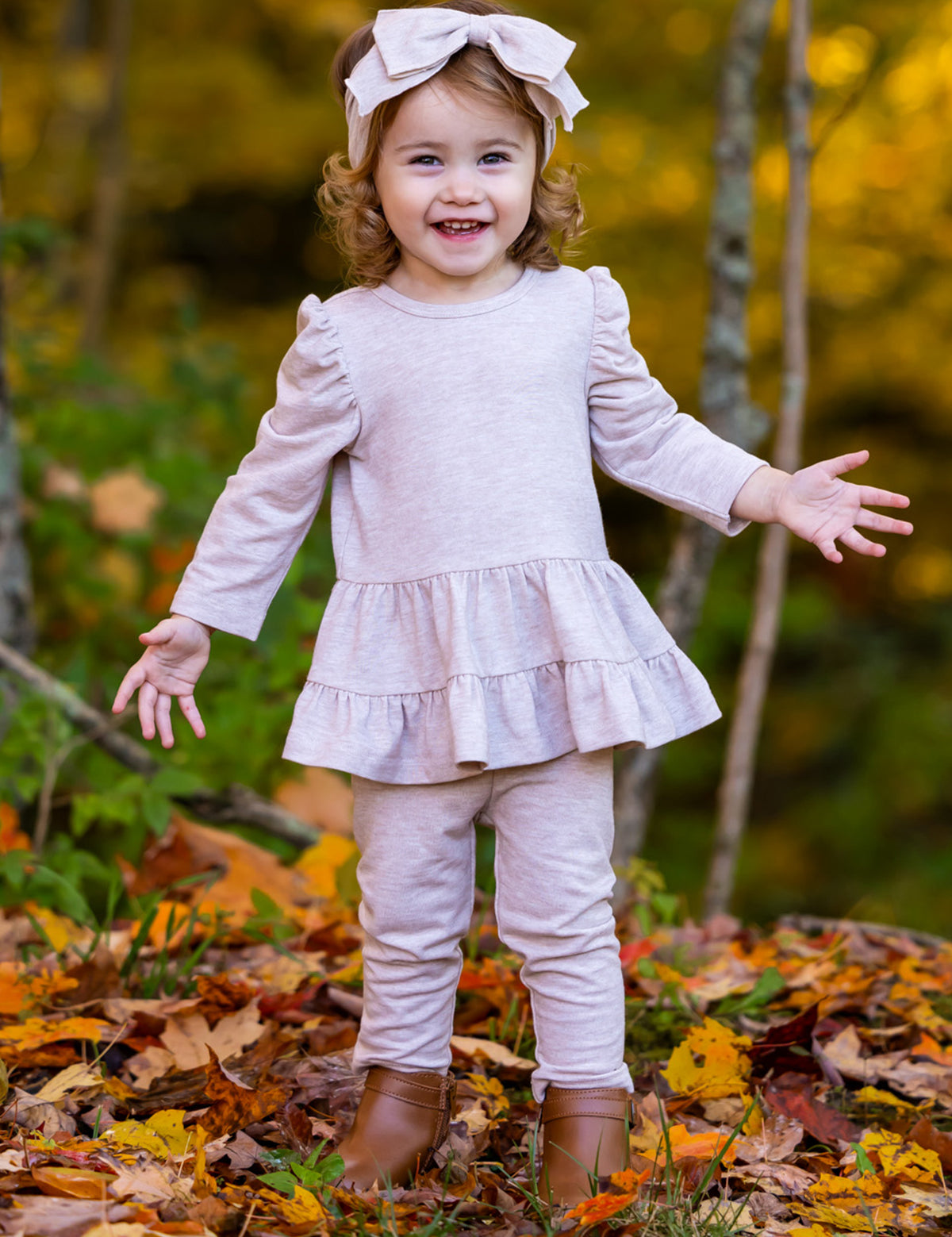 A smiling toddler with curly hair wears the Millie Two Piece Set by Mabel and Honey, paired with leggings, brown boots, and a pink bow headband. She stands outdoors on autumn leaves with trees in the background.