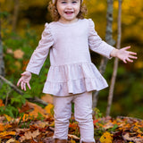 A smiling toddler with curly hair wears the Millie Two Piece Set by Mabel and Honey, paired with leggings, brown boots, and a pink bow headband. She stands outdoors on autumn leaves with trees in the background.