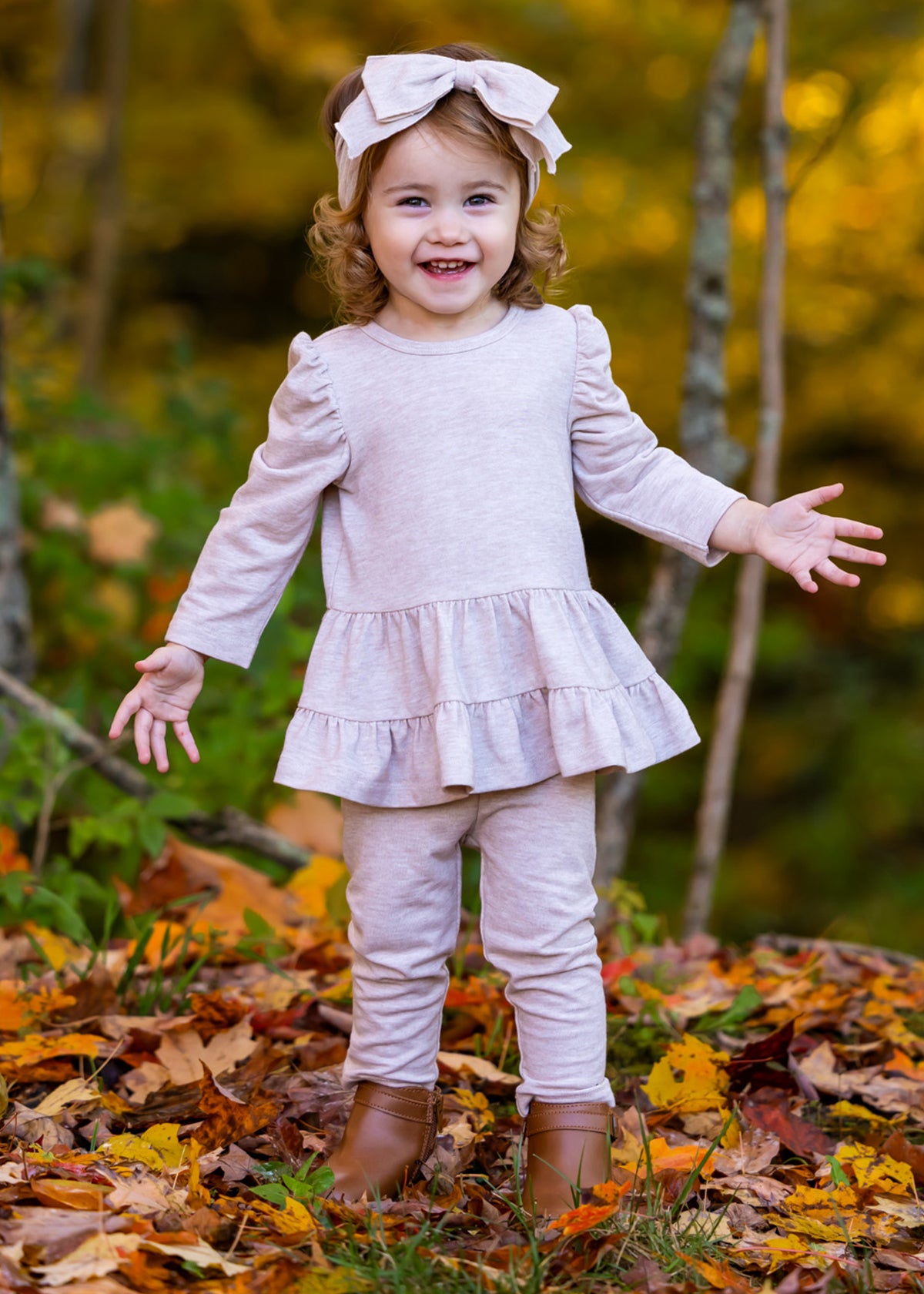A smiling toddler with curly hair wears the Millie Two Piece Set by Mabel and Honey, paired with leggings, brown boots, and a pink bow headband. She stands outdoors on autumn leaves with trees in the background.