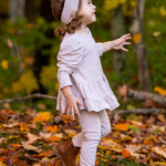 A young child wears the Millie Two Piece Set by Mabel and Honey, a light pink long sleeve outfit, paired with brown boots, walking through fallen autumn leaves with vibrant trees in the background.