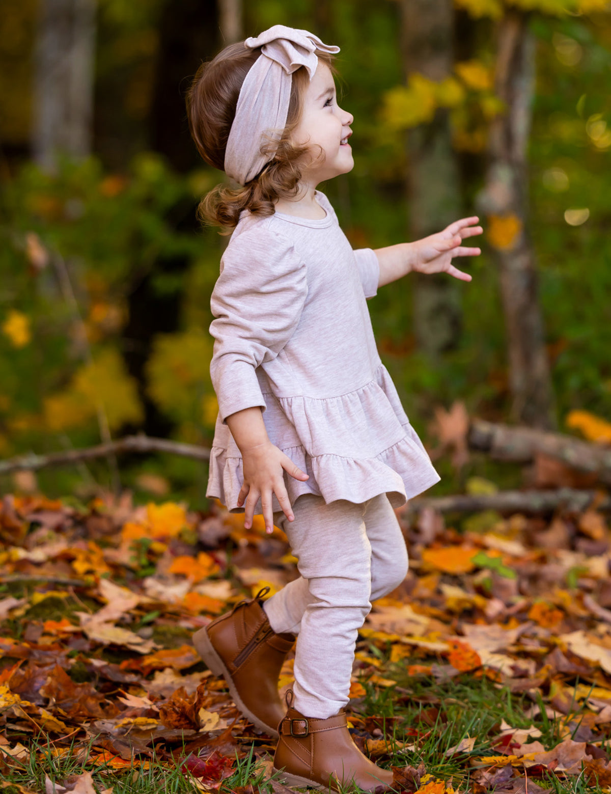 A young child wears the Millie Two Piece Set by Mabel and Honey, a light pink long sleeve outfit, paired with brown boots, walking through fallen autumn leaves with vibrant trees in the background.