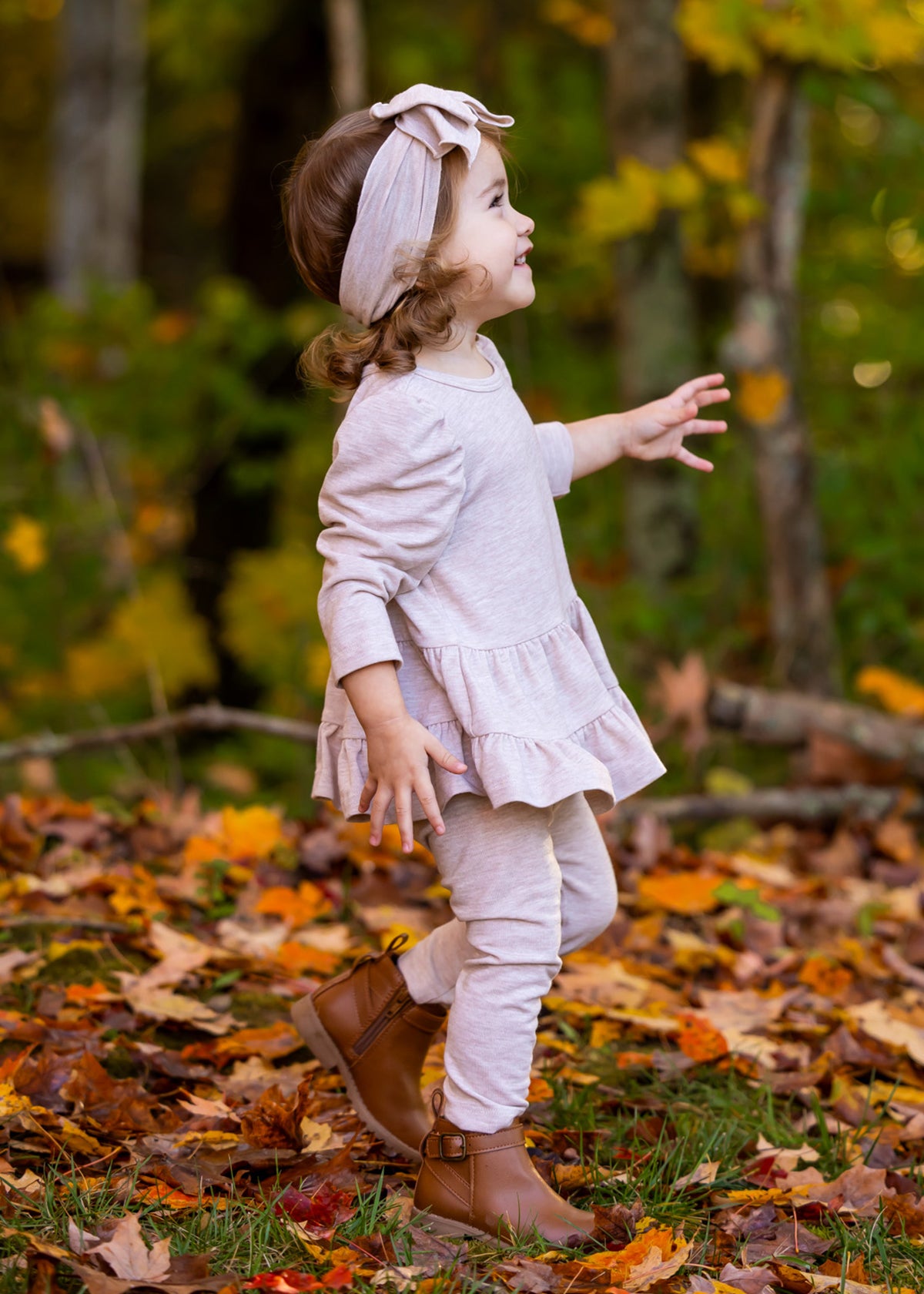 A young child wears the Millie Two Piece Set by Mabel and Honey, a light pink long sleeve outfit, paired with brown boots, walking through fallen autumn leaves with vibrant trees in the background.