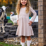 A young girl with curly blonde hair smiles in front of a wire fence with goats and a white chicken, wearing the Mabel and Honey Millie Natural Waist Dress, long socks, and brown shoes.