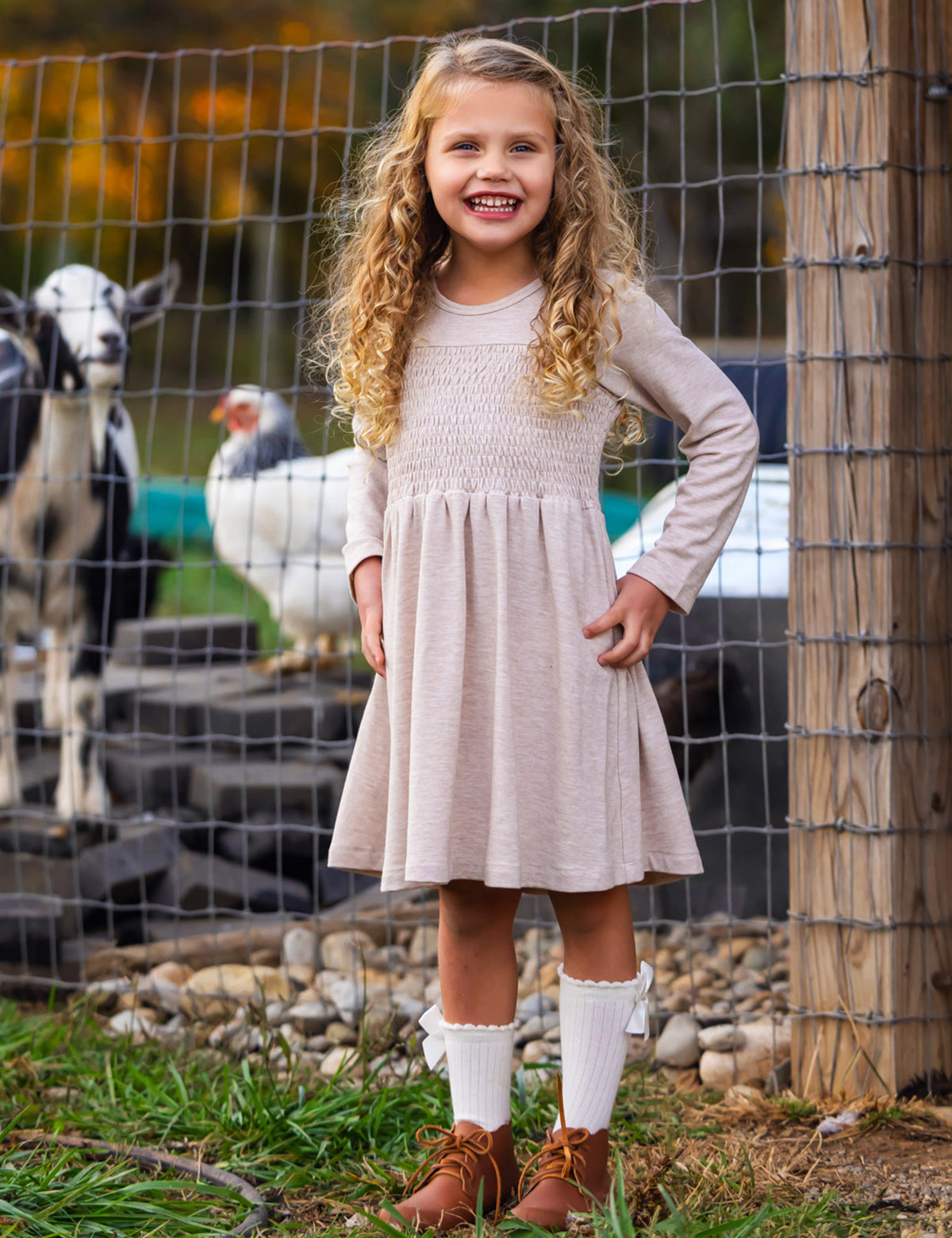 A young girl with curly blonde hair smiles in front of a wire fence with goats and a white chicken, wearing the Mabel and Honey Millie Natural Waist Dress, long socks, and brown shoes.
