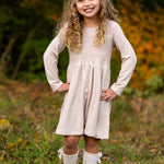 A smiling young girl stands on grass with her hands on her hips, wearing the Mabel and Honey Millie Natural Waist Dress, knee-high white socks with bows, and brown shoes. Autumn foliage is softly blurred in the background.