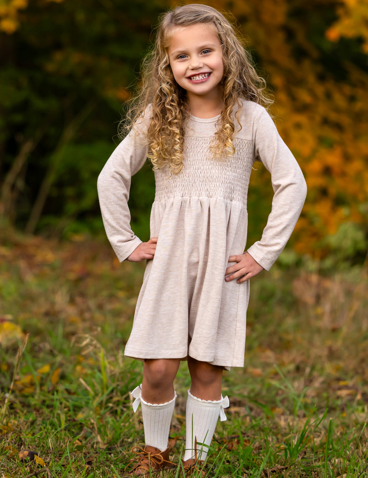 A smiling young girl stands on grass with her hands on her hips, wearing the Mabel and Honey Millie Natural Waist Dress, knee-high white socks with bows, and brown shoes. Autumn foliage is softly blurred in the background.