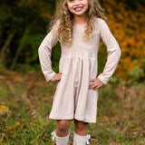 A smiling young girl stands on grass with her hands on her hips, wearing the Mabel and Honey Millie Natural Waist Dress, knee-high white socks with bows, and brown shoes. Autumn foliage is softly blurred in the background.