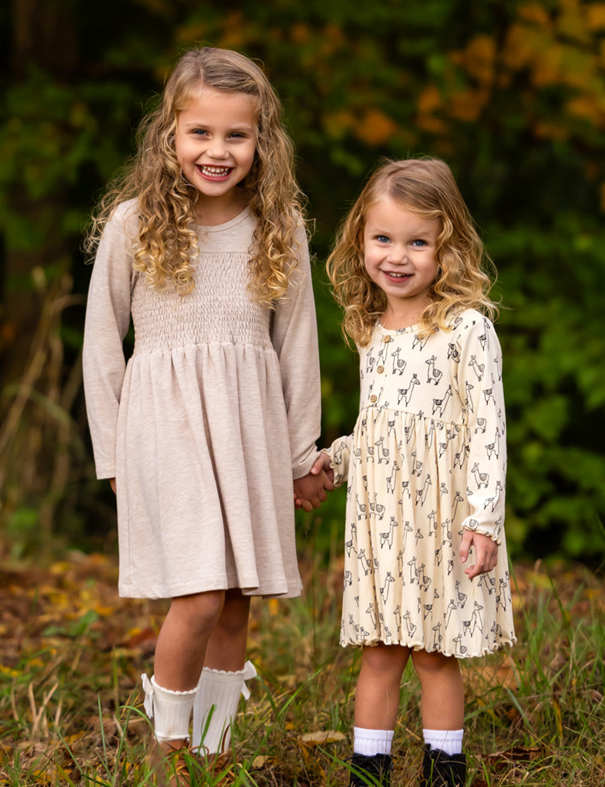 Two young girls with curly hair stand outdoors, holding hands and smiling. Both wear the Millie Natural Waist Dress from Mabel and Honey with ankle socks, surrounded by greenery and autumn foliage.