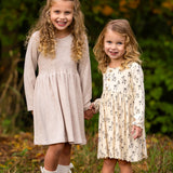 Two young girls with curly hair stand outdoors, holding hands and smiling. Both wear the Millie Natural Waist Dress from Mabel and Honey with ankle socks, surrounded by greenery and autumn foliage.