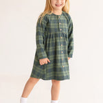 A young girl with long blonde hair smiles in the Cozy Creatures Dress by Mabel and Honey, featuring a green plaid pattern, long sleeves, and wooden buttons, paired with white socks and shoes on a plain light background.