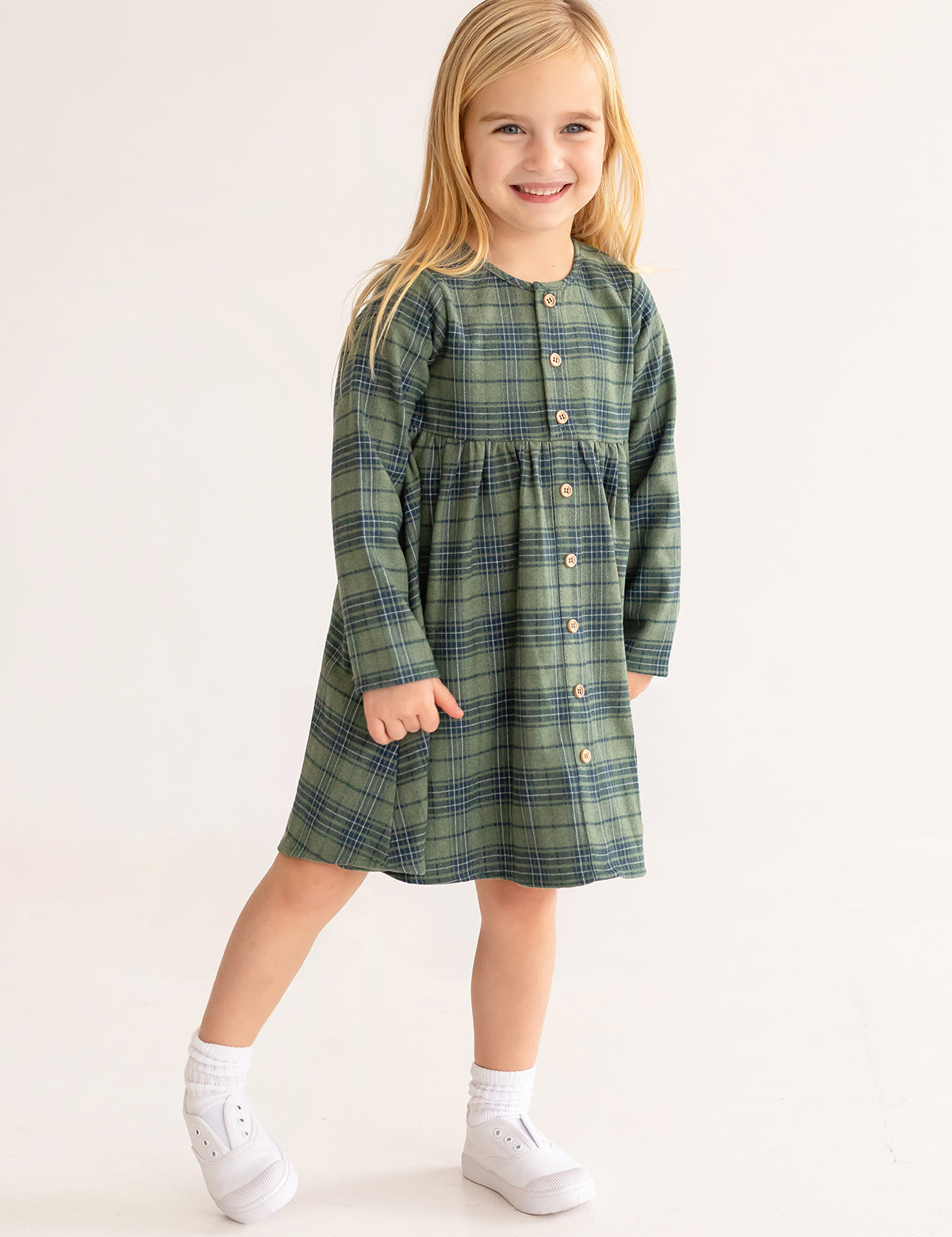 A young girl with long blonde hair smiles in the Cozy Creatures Dress by Mabel and Honey, featuring a green plaid pattern, long sleeves, and wooden buttons, paired with white socks and shoes on a plain light background.