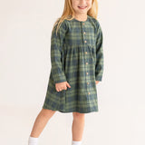 A young girl with long blonde hair smiles in the Cozy Creatures Dress by Mabel and Honey, featuring a green plaid pattern, long sleeves, and wooden buttons, paired with white socks and shoes on a plain light background.