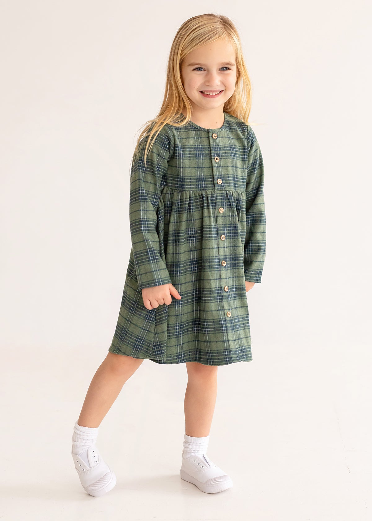 A young girl with long blonde hair smiles in the Cozy Creatures Dress by Mabel and Honey, featuring a green plaid pattern, long sleeves, and wooden buttons, paired with white socks and shoes on a plain light background.