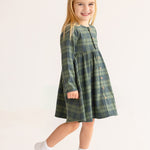 A young girl with long blonde hair smiles in the Cozy Creatures Dress by Mabel and Honey, featuring green plaid and wooden button details. She pairs it with white socks and sneakers, standing against a plain light background.