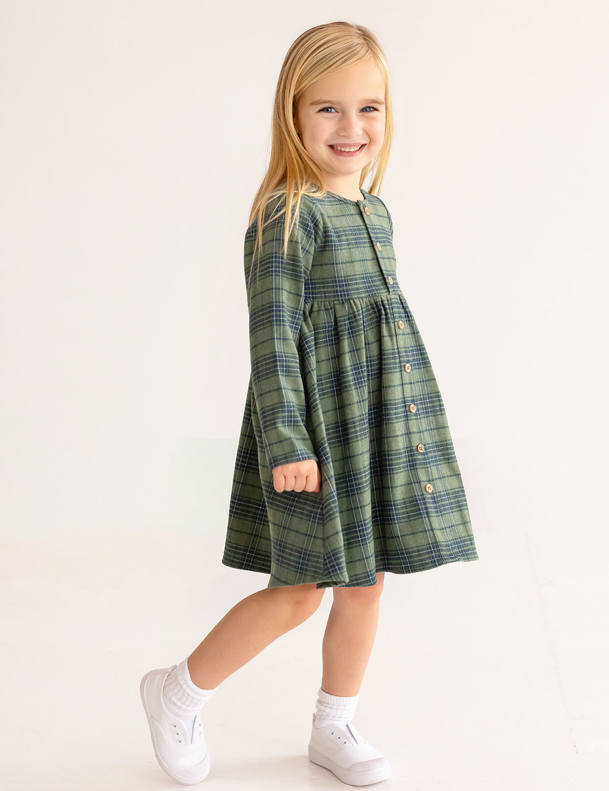 A young girl with long blonde hair smiles in the Cozy Creatures Dress by Mabel and Honey, featuring green plaid and wooden button details. She pairs it with white socks and sneakers, standing against a plain light background.