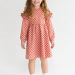 A young girl with long curly hair smiles while wearing the Mabel and Honey Red Velvet Dress, paired with white socks and sneakers, standing against a plain light background.