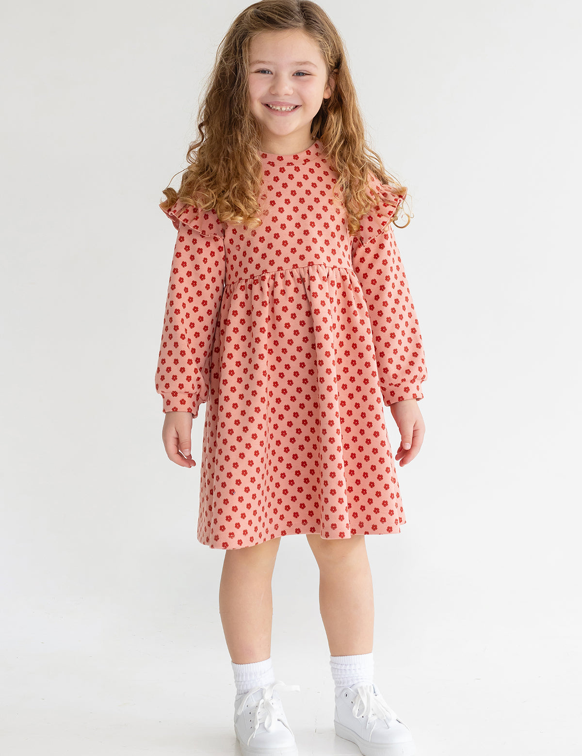 A young girl with long curly hair smiles while wearing the Mabel and Honey Red Velvet Dress, paired with white socks and sneakers, standing against a plain light background.