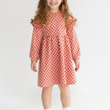 A young girl with long curly hair smiles while wearing the Mabel and Honey Red Velvet Dress, paired with white socks and sneakers, standing against a plain light background.