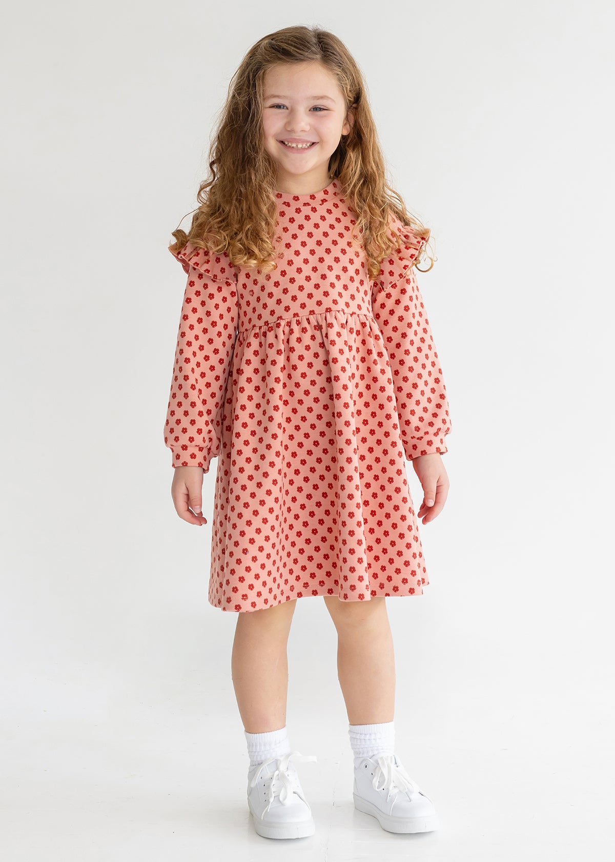 A young girl with long curly hair smiles while wearing the Mabel and Honey Red Velvet Dress, paired with white socks and sneakers, standing against a plain light background.