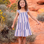 A young girl smiles outdoors on reddish-orange desert soil, surrounded by shrubs and yellow wildflowers, wearing the Riley Rose Dress by Mabel and Honey— a multicolor striped dress with ruffle shoulder straps and sandals.