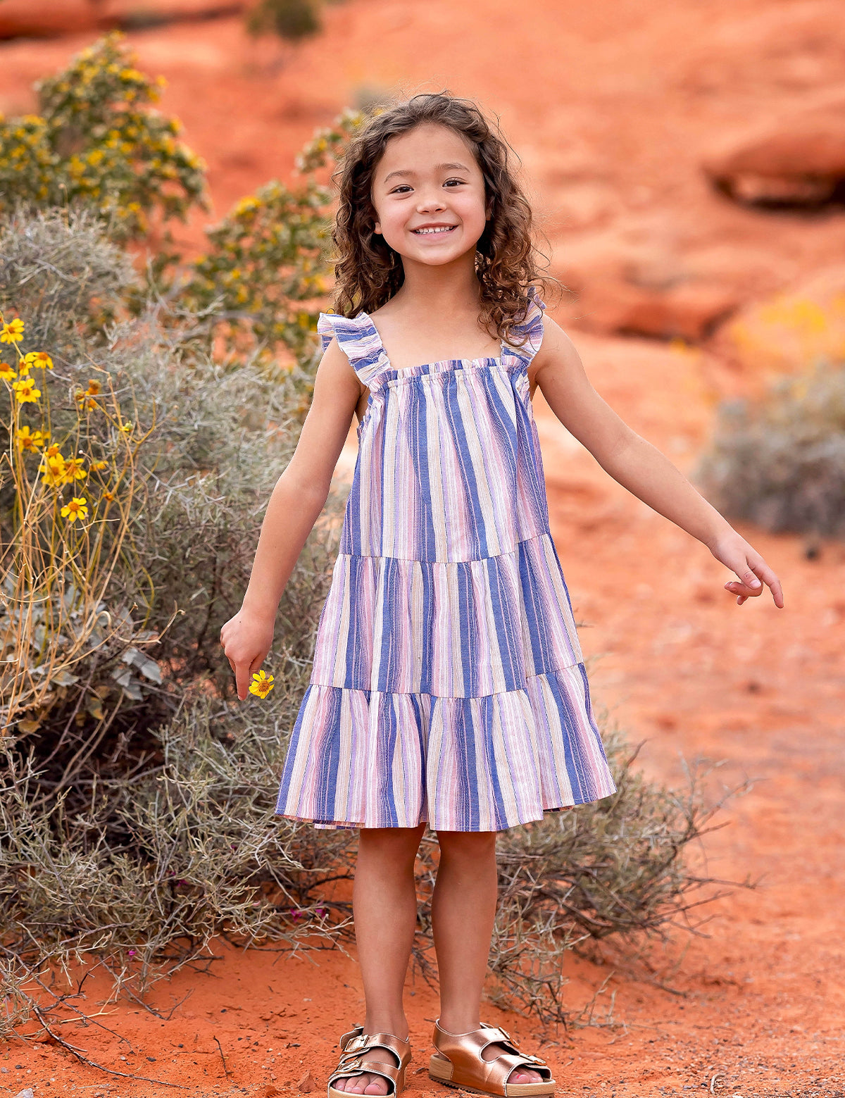 A young girl smiles outdoors on reddish-orange desert soil, surrounded by shrubs and yellow wildflowers, wearing the Riley Rose Dress by Mabel and Honey— a multicolor striped dress with ruffle shoulder straps and sandals.