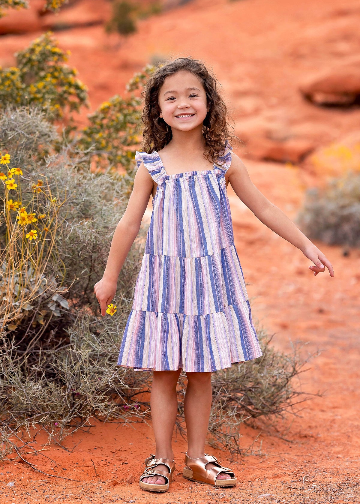 A young girl smiles outdoors on reddish-orange desert soil, surrounded by shrubs and yellow wildflowers, wearing the Riley Rose Dress by Mabel and Honey— a multicolor striped dress with ruffle shoulder straps and sandals.
