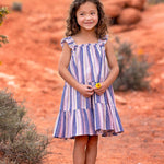 A young girl with curly hair smiles outdoors on red dirt, wearing the Riley Rose Dress by Mabel and Honey. She holds a small yellow flower, with bushes and red rocks in the background.
