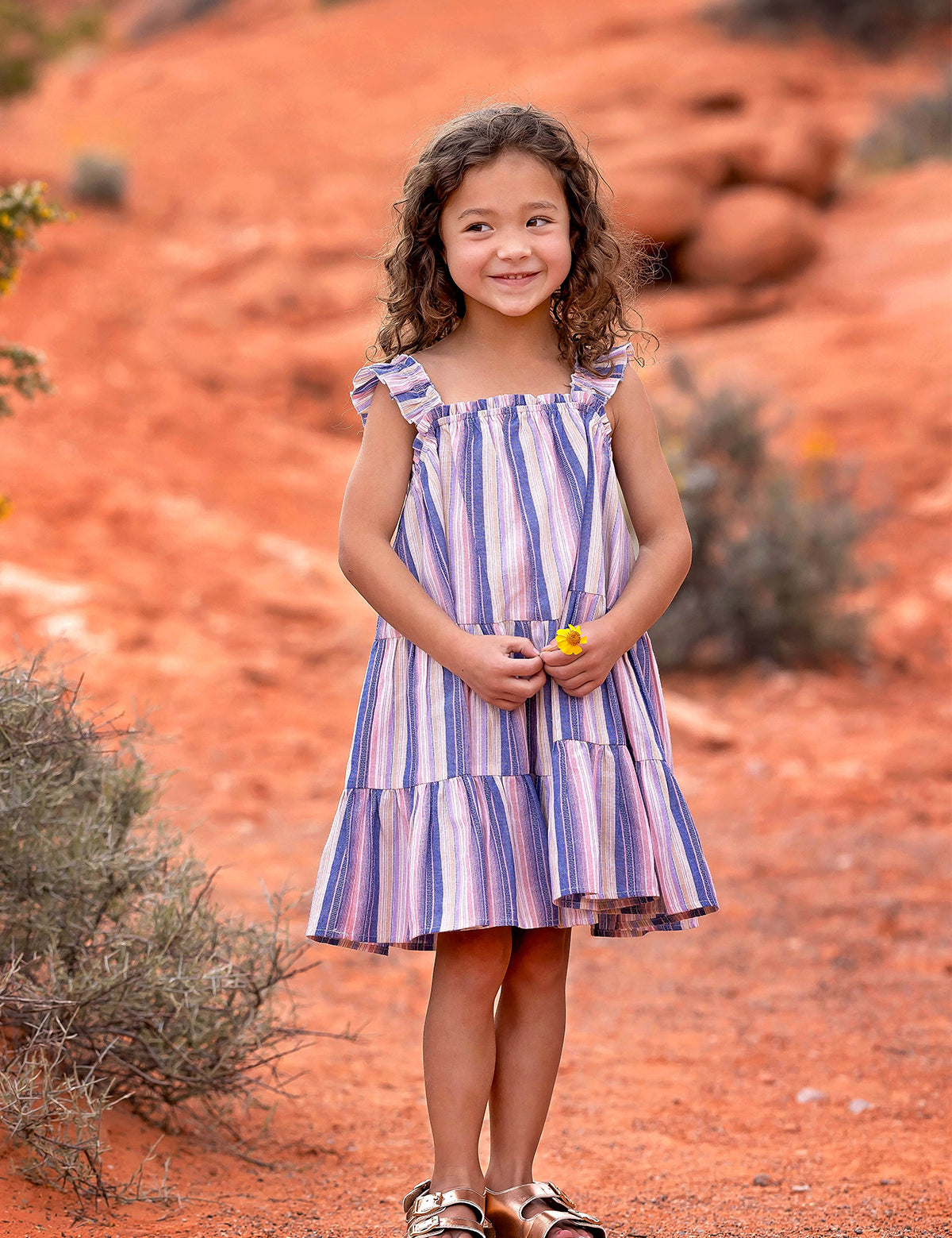 A young girl with curly hair smiles outdoors on red dirt, wearing the Riley Rose Dress by Mabel and Honey. She holds a small yellow flower, with bushes and red rocks in the background.