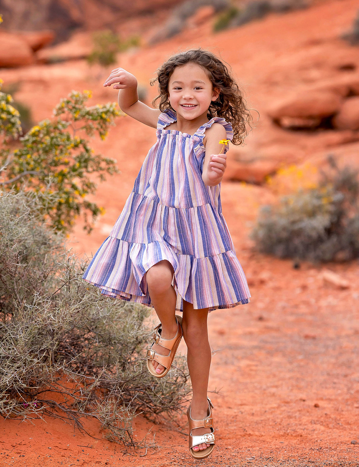 A young girl with curly hair joyfully jumps outdoors in the Riley Rose Dress by Mabel and Honey, featuring multicolor stripes and ruffle shoulder straps, against a red sandy landscape with rocks and shrubs in the background.