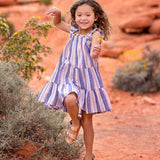 A young girl with curly hair joyfully jumps outdoors in the Riley Rose Dress by Mabel and Honey, featuring multicolor stripes and ruffle shoulder straps, against a red sandy landscape with rocks and shrubs in the background.