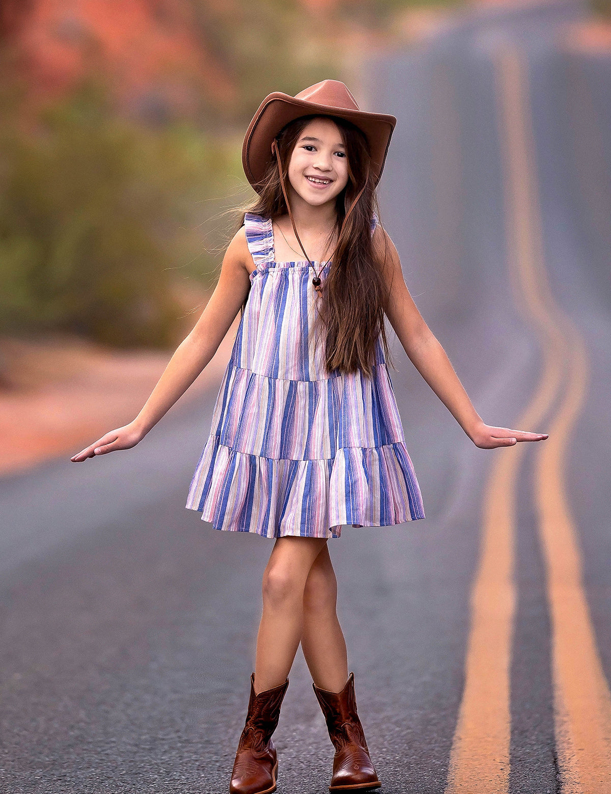 A young girl wears the Mabel and Honey Riley Rose Dress with colorful stripes and ruffle straps, smiling on a winding desert road in brown cowboy boots and a wide-brimmed hat.