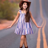 A young girl wears the Mabel and Honey Riley Rose Dress with colorful stripes and ruffle straps, smiling on a winding desert road in brown cowboy boots and a wide-brimmed hat.
